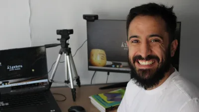 Juani sonriendo en su escritorio, con la cámara y la pantalla desde donde graba y da sus clases
